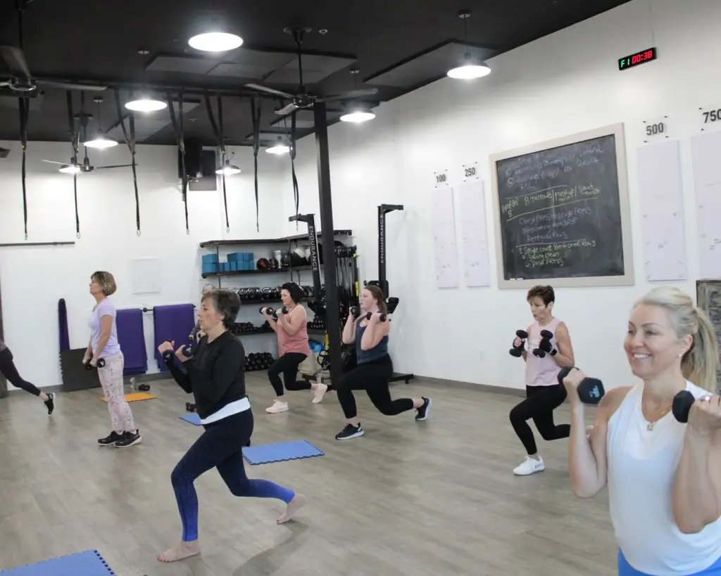 Clemson Fitness class, studio full of women, bending right leg while lifting dumbbells in both hands. Smiling and having fun.